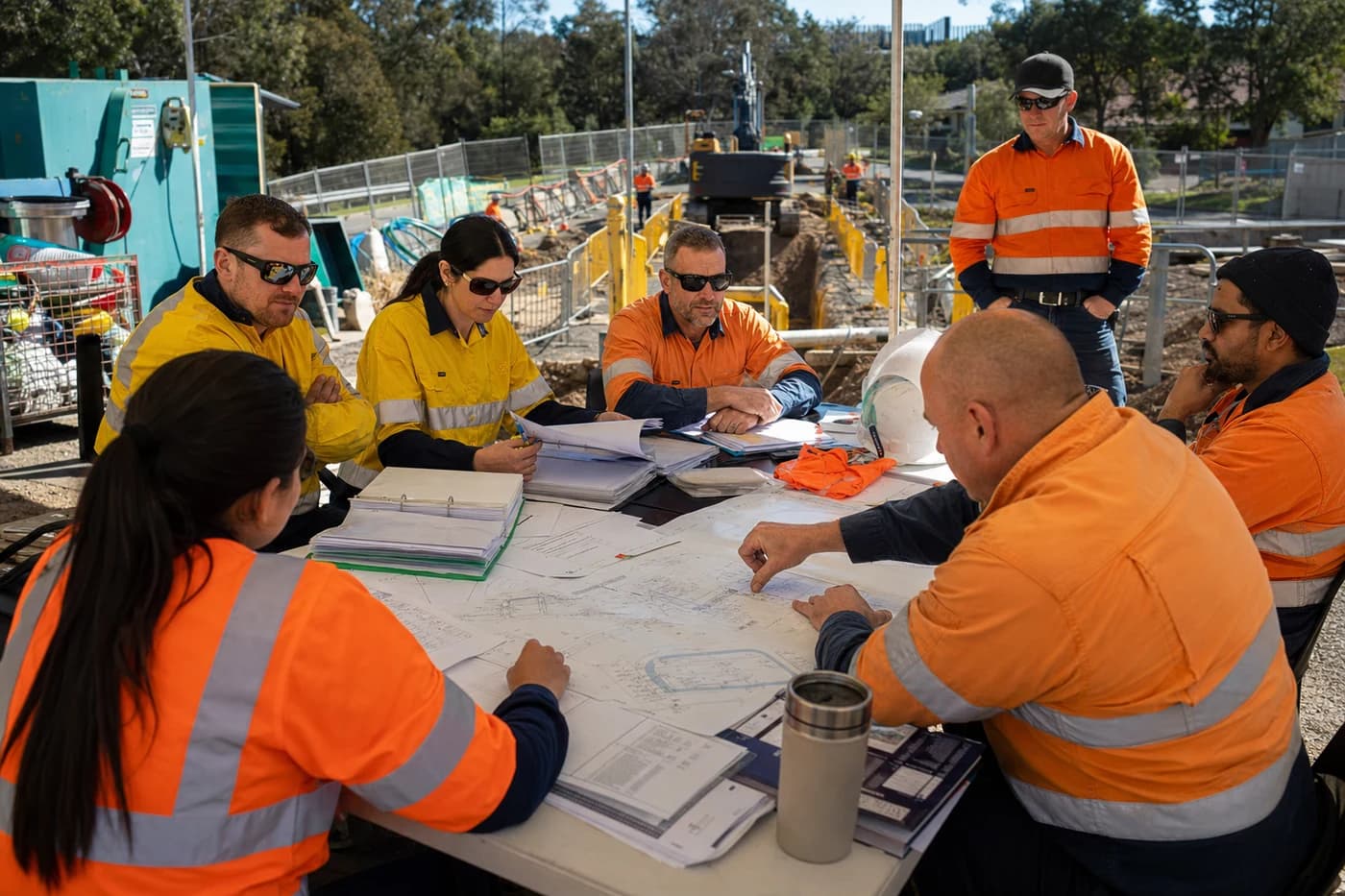 Live water network construction pre-start meeting at a Sydney site compound with project team reviewing plans, SWMS folders, and isolation diagrams
