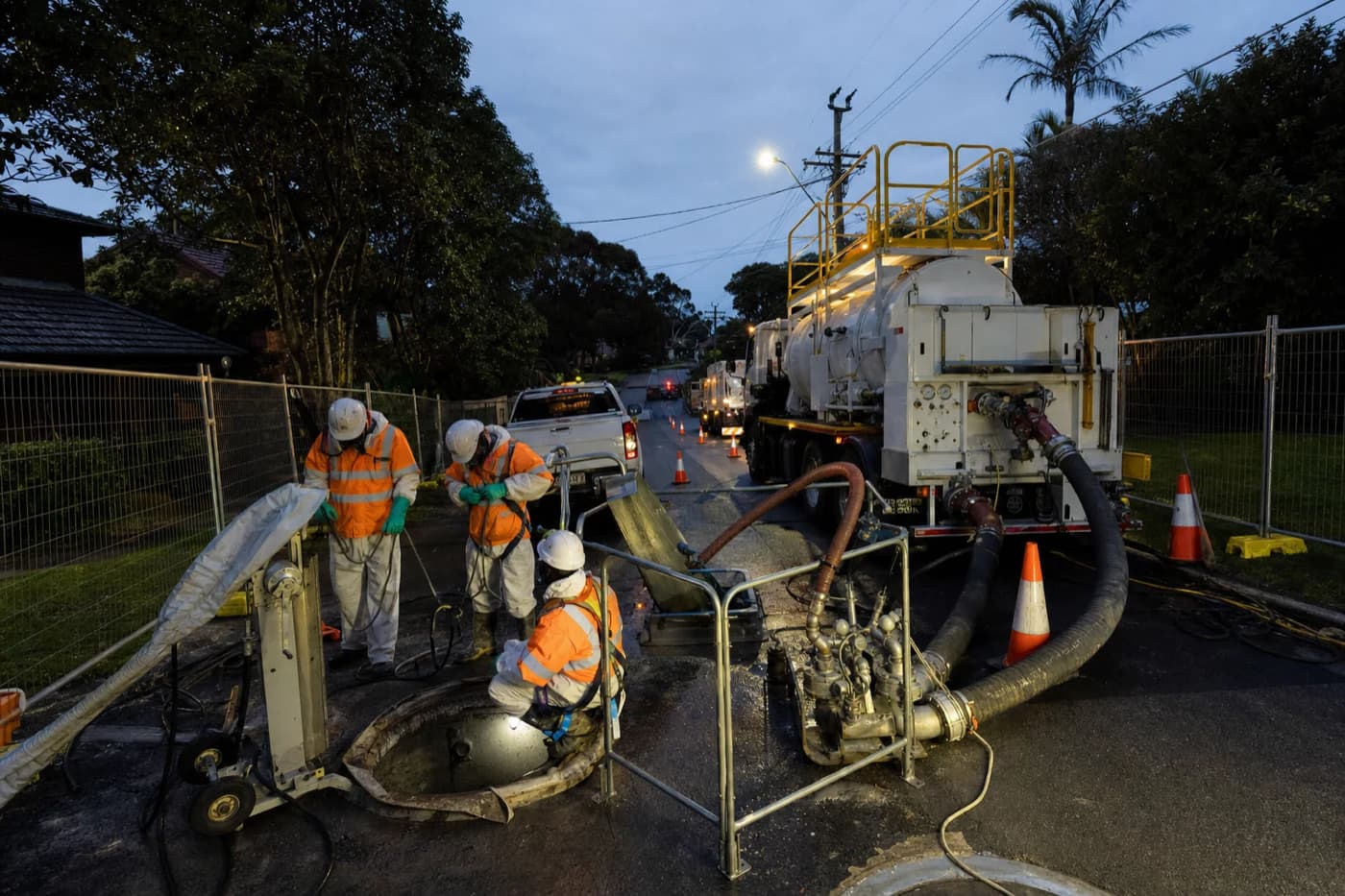 Wastewater rehabilitation site at dusk on a Sydney suburban street with manhole access, bypass pumping, and crew in hygiene PPE